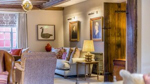 Uma sala de estar aconchegante com um sofá, poltrona e uma mesa com abajur. Há quadros nas paredes e uma porta de madeira.