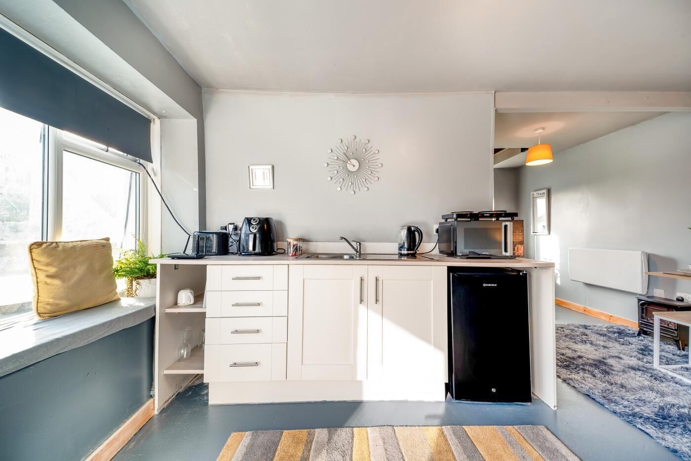 A modern kitchen with white cabinets, a built-in dishwasher, and a microwave. There is a window providing natural light and a decorative wall clock.
