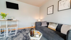 Una sala de estar moderna con un sofá gris, una mesa de centro con tetera y taza, una pequeña mesa de comedor con sillas y un televisor de pantalla plana montado en la pared.