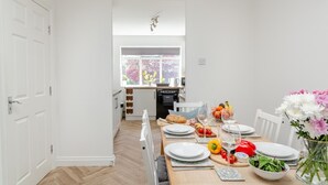 Uma cozinha moderna com armários brancos, uma mesa de jantar posta para quatro pessoas, com pratos, copos e um vaso de flores.