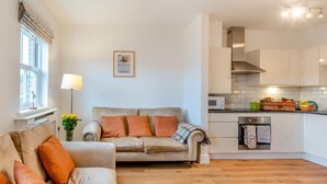 Un salon moderne avec un canapé beige et des coussins orange, une petite table avec une lampe, et un coin cuisine doté de meubles blancs et d’un four à micro-ondes.