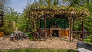 An outdoor seating area with a brick oven, wooden pergola, and a table with chairs.