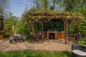 An outdoor seating area with a brick oven, wooden pergola, and a table with chairs.