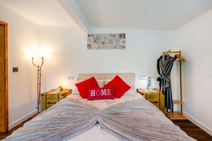 A bedroom with a bed, a bedside table, a lamp, a robe, a coat rack, and a painting on the wall.