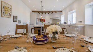 Un comedor con una mesa de madera dispuesta para seis personas, un jarrón con flores blancas y una cocina al fondo.