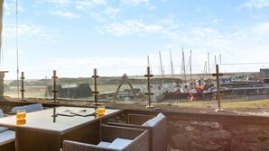 Un área de estar al aire libre con mobiliario de mimbre y una mesa, con vistas a una marina con barcos y un puente.