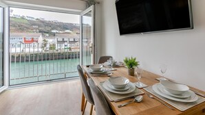 A dining area with a view of a river and houses, a flat-screen TV on the wall, and a wooden table set for four. - 3 Bed in Portreath (oc-c30852) (Portreath)