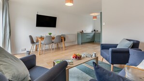A modern living room with a sofa set, a coffee table with magazines and fruit, a dining area with chairs and a table, and a kitchen with cabinets and a TV mounted on the wall. - 3 Bed in Portreath (oc-c30852) (Portreath)