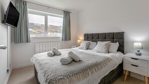 A neatly made bed with pillows and a blanket, a bedside table with a lamp, a TV mounted on the wall, and a window with curtains. - 3 Bed in Portreath (oc-c30852) (Portreath)
