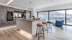 A modern kitchen with a central island, bar stools, and a view of a residential area. - 5 Bed in Mortehoe (oc-c30855) (Mortehoe)