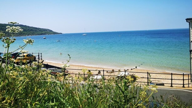A coastal view with a clear blue sea, a sandy beach, and a few boats. There is a building on the right side of the image.