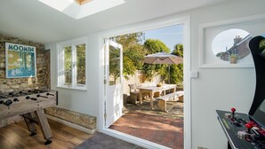 A room with a foosball table, a gaming console, a dining area with a table and chairs, and a view of the garden through a glass door.