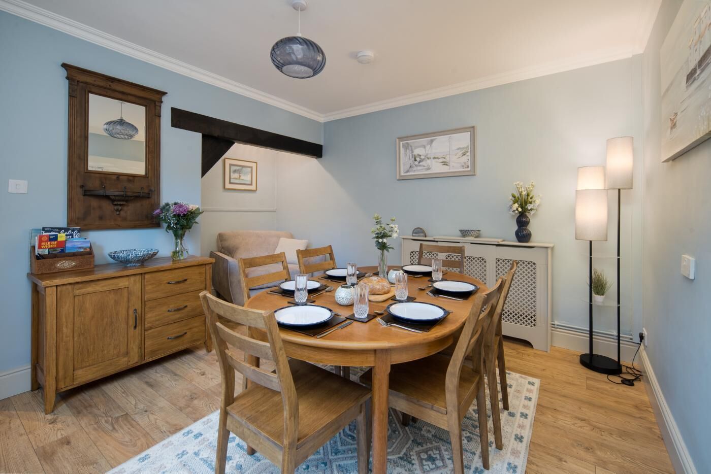 Uma sala de jantar com uma mesa de madeira posta para quatro pessoas, rodeada por cadeiras de madeira. Um aparador de madeira com gavetas fica à esquerda, e uma pequena mesa lateral com um vaso de flores à direita.