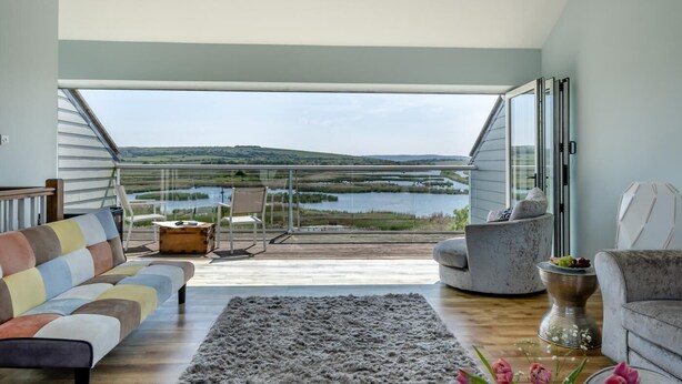 A spacious living room with a large window overlooking a lake and hills. There is a colorful sofa, a grey rug, and a round table with a decorative vase. - 3 Bed in St. Helens (oc-y30961) (Saint Helens)