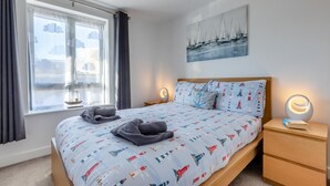 Una cama bien hecha con una colcha de temática náutica, una lámpara de noche y vistas al mar a través de una ventana con cortinas.