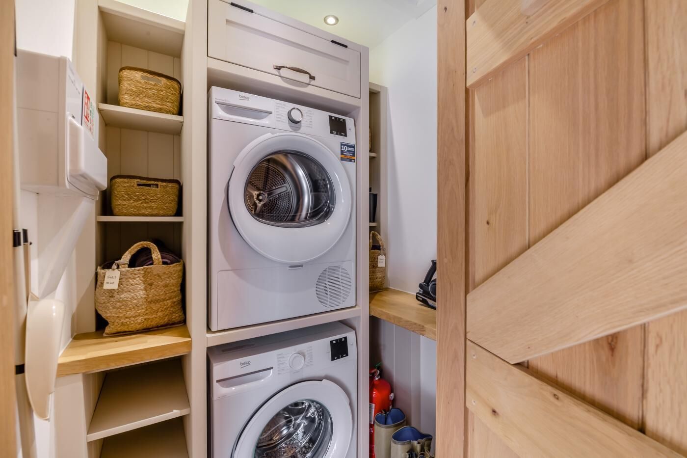 A laundry area with two front-loading washing machines, wooden shelves with baskets, and a fire extinguisher.