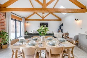 Un coin repas avec une table en bois dressée pour six personnes, un canapé en arrière-plan et un mur en briques.