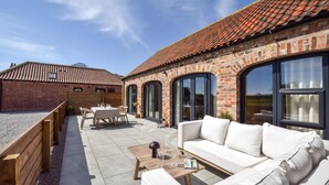 A patio area with a white sofa, a wooden table, and a brick building with large windows. - 2 Bed in Beeford (oc-t90892) (Beeford)