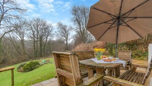 En udendørs træterrasse med et bord, stole og en parasol, samt udsigt til en have.