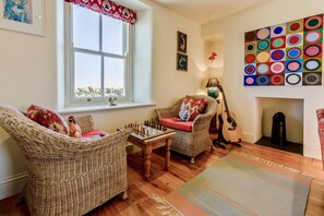 A cozy living room with wicker furniture, a chessboard, a guitar, and a colorful wall art piece. - 3 Bed in Penzance (oc-k20665) (Lamorna)
