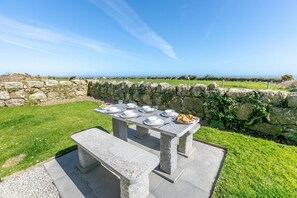 A stone table set outdoors with plates, glasses, and food, overlooking a grassy area and stone wall. - 3 Bed in Penzance (oc-k20665) (Lamorna)