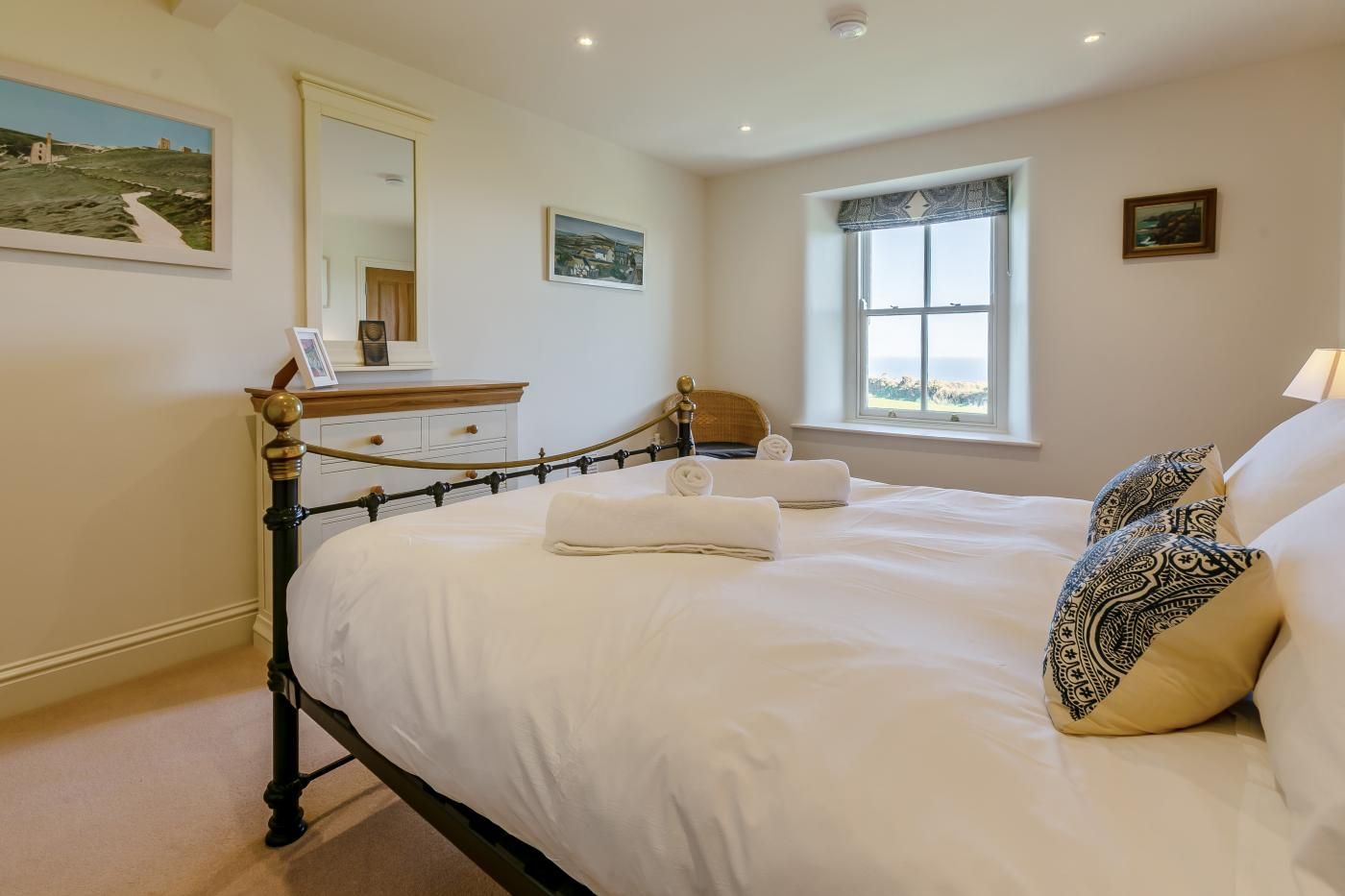 Een netjes opgemaakt bed met witte lakens en een hoofdbord, een nachtkastje met een lamp, en een raam met uitzicht.