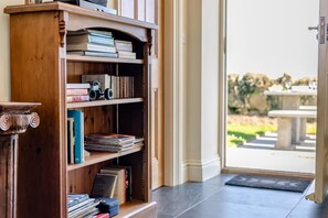 Una estantería de madera llena de libros y un coche de juguete, una puerta que da al exterior, y un banco de piedra al fondo.