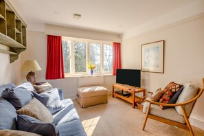 Un salon comprenant un canapé, une table basse en bois, un téléviseur, un fauteuil, une lampe et une fenêtre avec des rideaux.