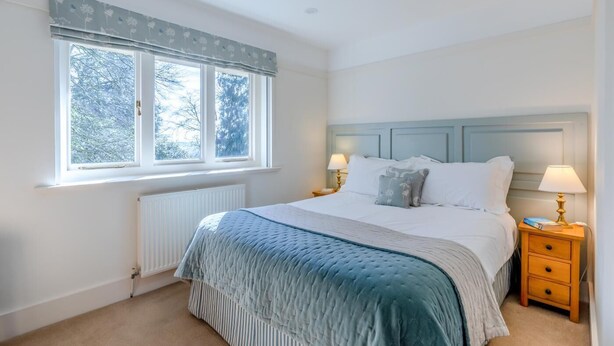 A bedroom with a bed, two bedside tables, a window with curtains, and a radiator. - 2 Bed in East Anstey (oc-k31022) (East Anstey)