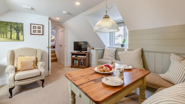 A cozy living room with a wooden coffee table, striped armchair, and a sofa. A television and cabinet are in the background. - 1 Bed in East Anstey (oc-w31023) (East Anstey)