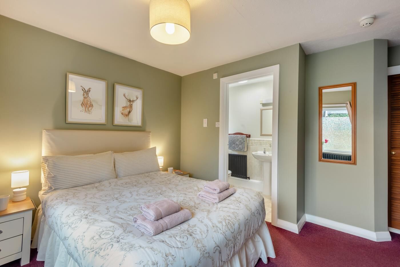 Ein Schlafzimmer mit einem Bett, zwei Nachttischlampen, einem Nachttisch, einem Bild von einem Reh im Bilderrahmen, einem durchgehenden Türspalt sichtbaren Badezimmer und einem Spiegel an der Wand.