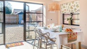 Un comedor con una mesa dispuesta para cuatro, una lámpara colgante de mimbre y vistas a un patio trasero con cerca de madera y edificios de ladrillo.