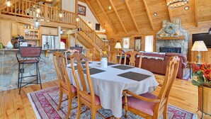 A spacious living area with a high ceiling, wooden beams, a stone fireplace, and a dining table set for four. - 1 Mi to Lake Tillery: Outdoor Retreat w/ Hot Tub! (Mt Gilead)