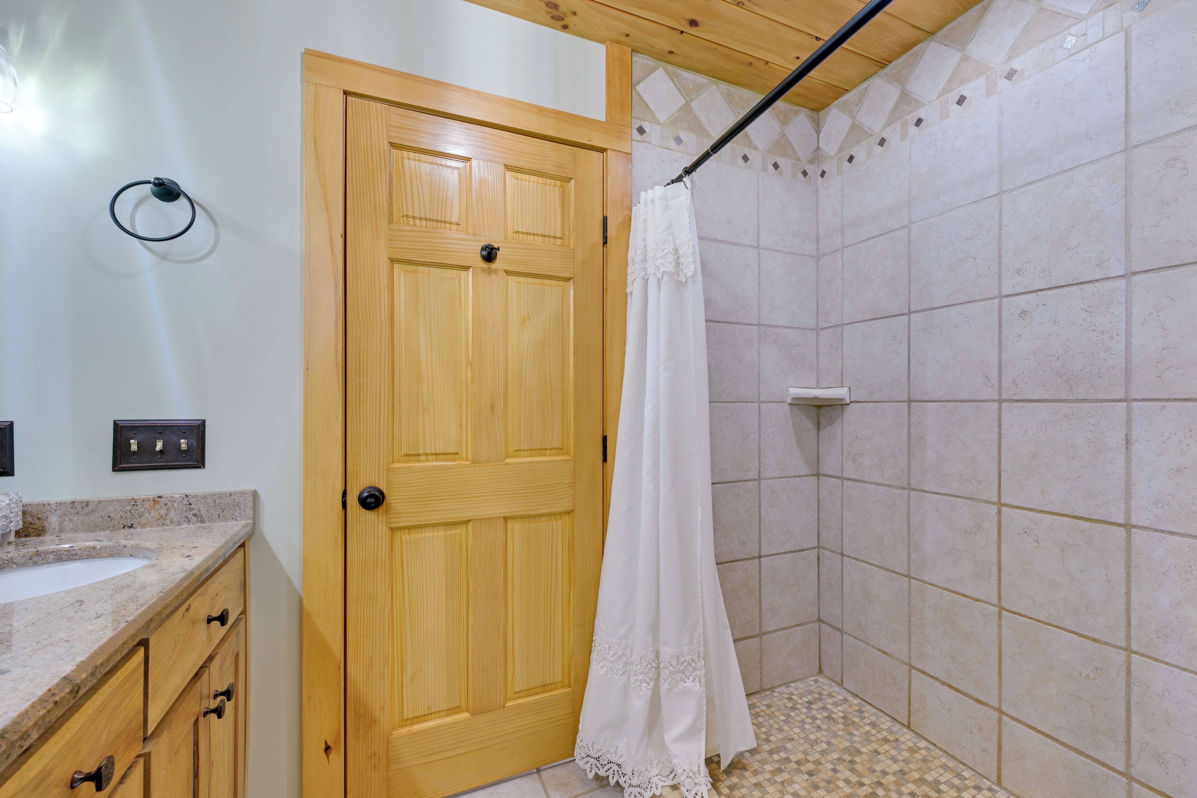 Une salle de bain avec une porte en bois, une douche avec un rideau blanc, un lavabo avec un comptoir en granit et un sol de douche carrelé.