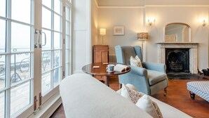 A room with a large window, a fireplace, a sofa, a chair, and a table with a book and a cup. - 7 Bed in Tenby (oc-b30996) (Tenby)