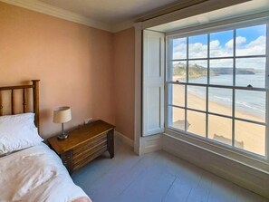A bedroom with a bed, a nightstand, a lamp, and a large window overlooking a beach. - 7 Bed in Tenby (oc-b30996) (Tenby)