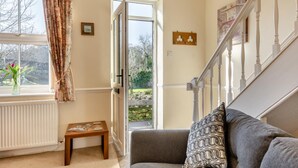 A cozy living room with a sofa, a small table, a window with curtains, and a view of a garden through an open door.