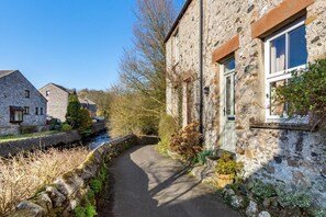 A narrow stone pathway leading to a stone building with a window and a door, surrounded by greenery and a small stream. - 2 Bed in Ingleton (oc-w31066) (Ingleton)