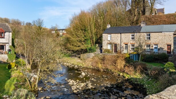 A stone building with a river in front, surrounded by trees and a clear sky.