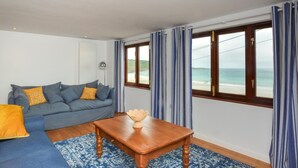 Een kamer met een blauwe bank, een houten tafel en uitzicht op zee door grote ramen.