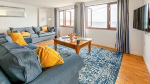 Een moderne woonkamer met een grijze bank, gele kussens, een houten salontafel en een vloerkleed met een patroon.