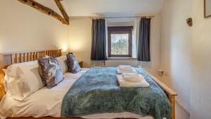 Ein Schlafzimmer mit einem Holzkopfteil, einem großen Bett mit weißer Bettwäsche, einer flauschigen, grünen Decke und zwei Handtüchern. Im Hintergrund ist ein Fenster mit Vorhängen zu sehen.