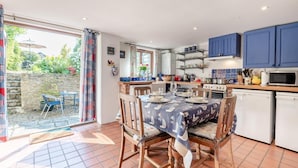 Une cuisine avec un coin repas, dotée d’un sol carrelé, de chaises en bois, d’une table dressée pour quatre personnes et de meubles de rangement bleus.