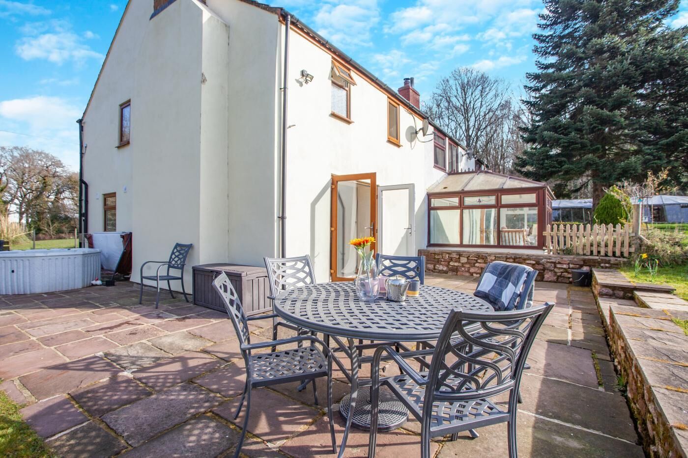 A white house with a patio featuring a round metal table and chairs, a wooden deck, and a stone wall.