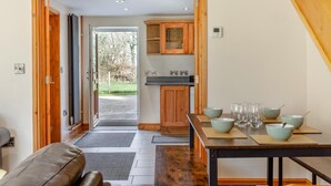 A cozy kitchen with wooden cabinets, a dining table with bowls and glasses, and a view of the outdoors through a glass door. - 3 Bed in Littledean (oc-g31084) (Littledean)