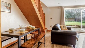 A modern living room with a wooden staircase, a dining table set with bowls and glasses, and a leather sofa with a cushion. - 3 Bed in Littledean (oc-g31084) (Littledean)