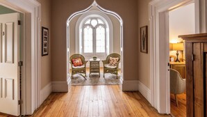 A hallway with a wooden floor, two wicker chairs with cushions, a small round table, and a large arched window with multiple panes. - Strathmere Country Retreat (Ottawa)
