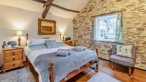 Quarto com parede de pedra, cama de madeira, uma cadeira e janela com cortinas.