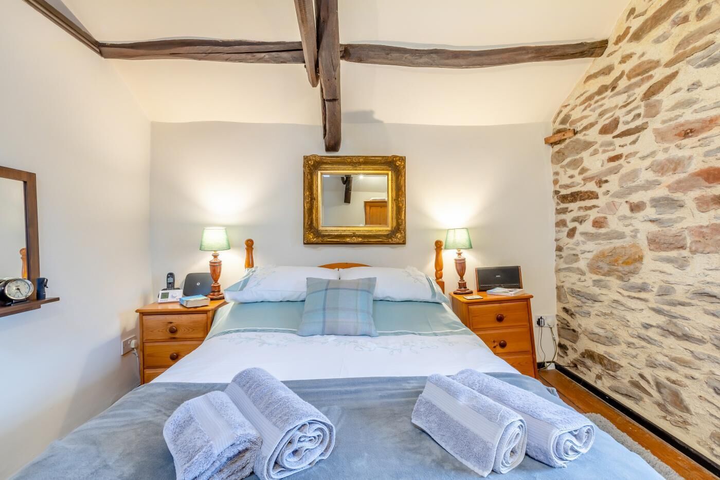 Une chambre à coucher avec un lit, deux tables de chevet, une commode, un miroir et un mur en pierre.