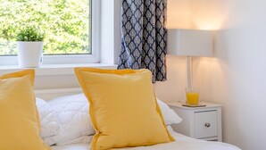 Ein ordentlich bezogenes Bett mit weißer Bettwäsche und zwei gelben Kissen, ein Nachttisch mit einem Glas Orangensaft, ein Topf mit einer Pflanze und ein gemustertes Vorhang.